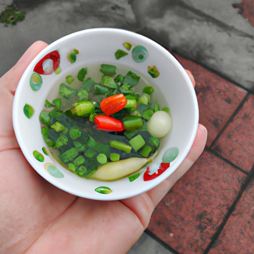 Một bàn tay đang cầm một chiếc bát nhỏ đựng đầy ốc nấu chuối xanh. Món ăn được trang trí với hành tây xanh và ớt chuông đỏ.